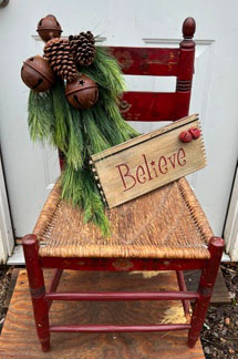 Antique woven seat chair painted red with Believe sign and greens with rusty bells on back.