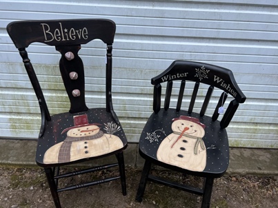 Two wooden chairs painted black first with the word believe on the back and snowman on the seat. The second chair has Winter Wishes painted on the backrest and a snowman and snowflake on the seat.