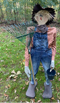 Tall wooden scarecrow dressed in overalls and shirt holding a rake