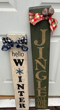 Two wooden signs. Left: Hellow winter is ivory background with black lettering and black and white bow. Right: Green sign with Old Ivory printing that says Jingle with plaid ribbon at the top.
