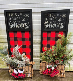 Two black wooden boards stenciled in white with This House Believes, red and black plaid background behind a wire basket filled with seasonal greenery and boots.