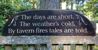 Black, wooden signed with stenciled lettering that says" The days are short, the weather's cold, by tavern fires tales are told."