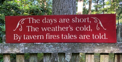 Red, wooden sign with stenciling that says "The days are short, the weather's cold, by tavern fires, tales are told." 